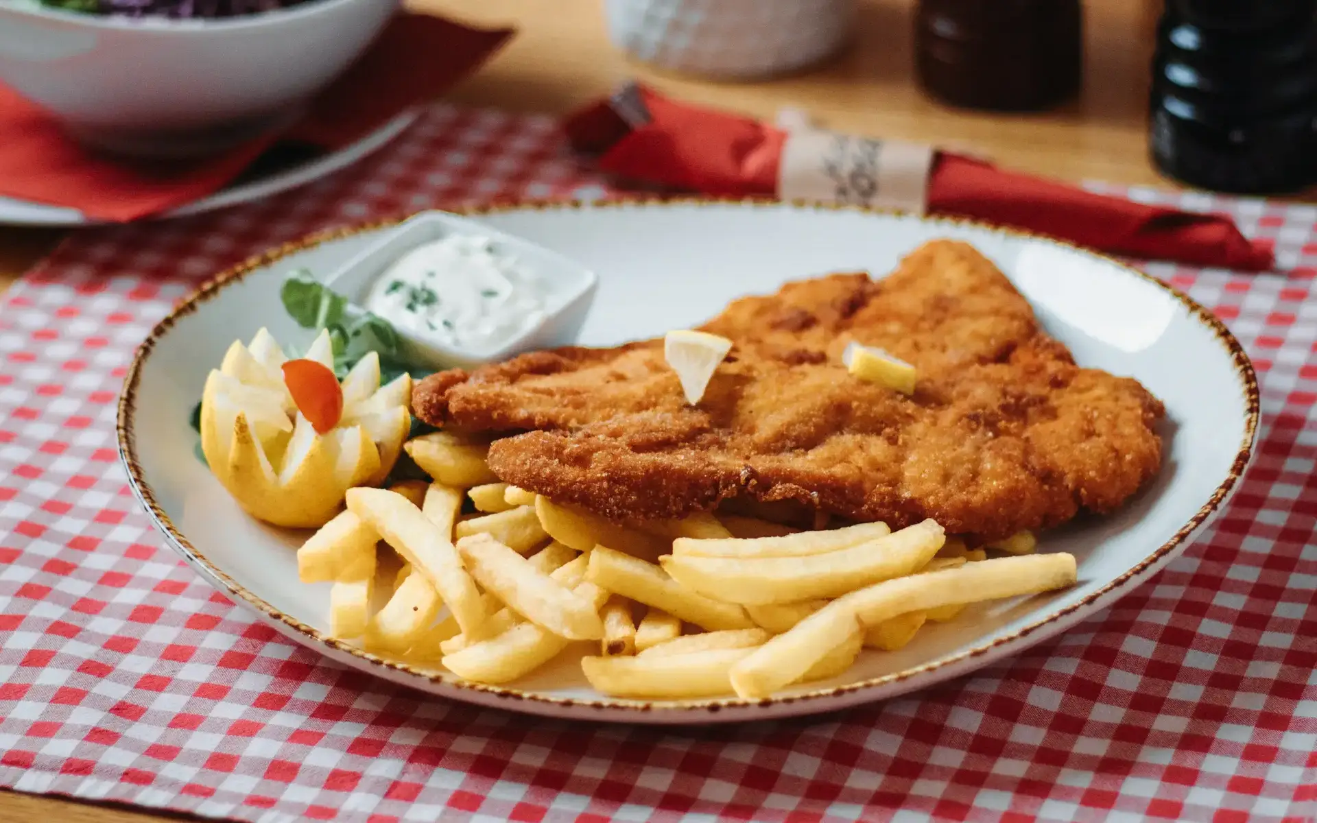 U perspektivi fotografiran tanjur sa Bečkim odreskom, lađicama krumpira, tartar umakom a sve skupa na crvenom pepita stolnjaku