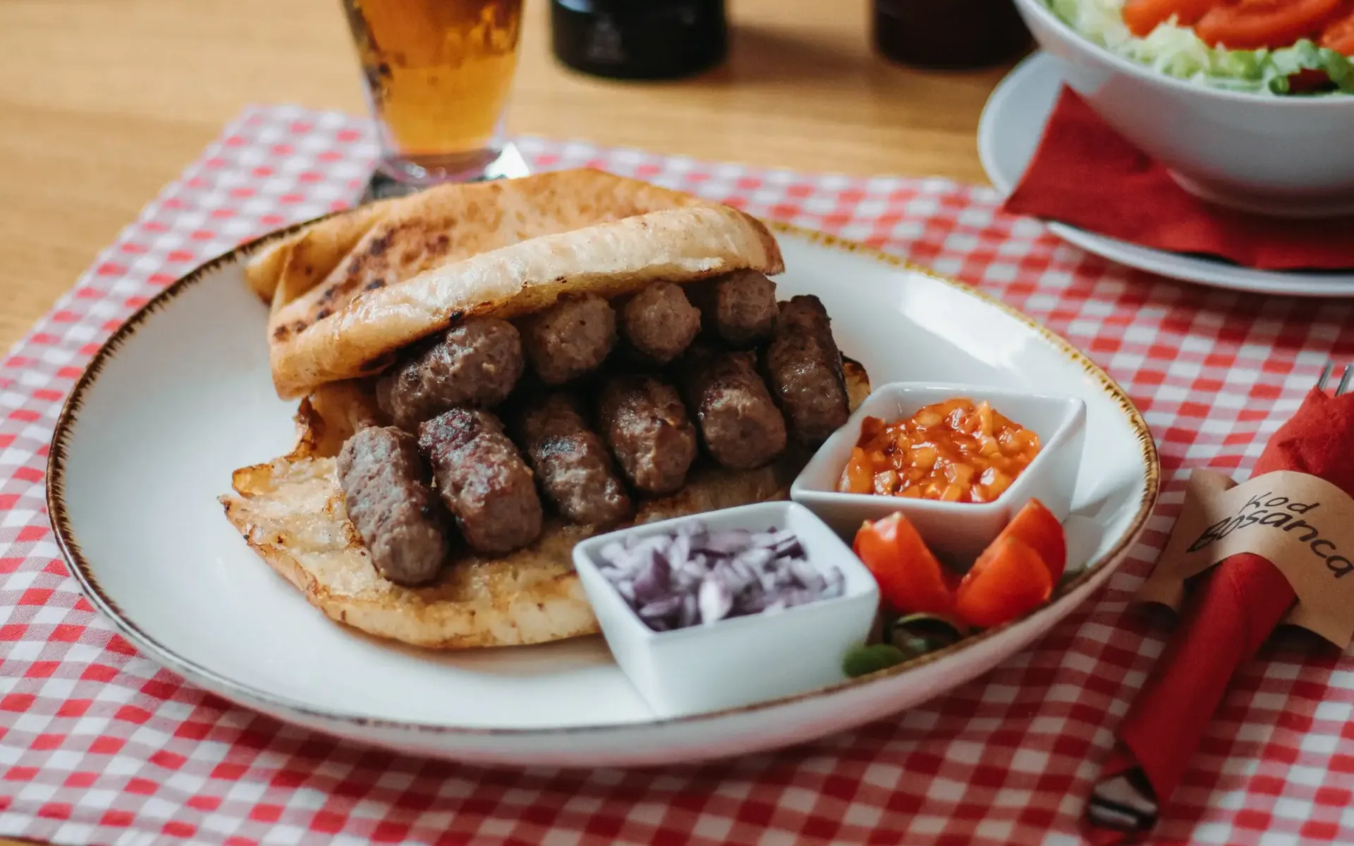 U perspektivi fotografiran tanjur sa Gurmanskoim pljeskavicom, ljubičastim lukom i roštilj umakom a sve skupa na crvenom pepita stolnjaku