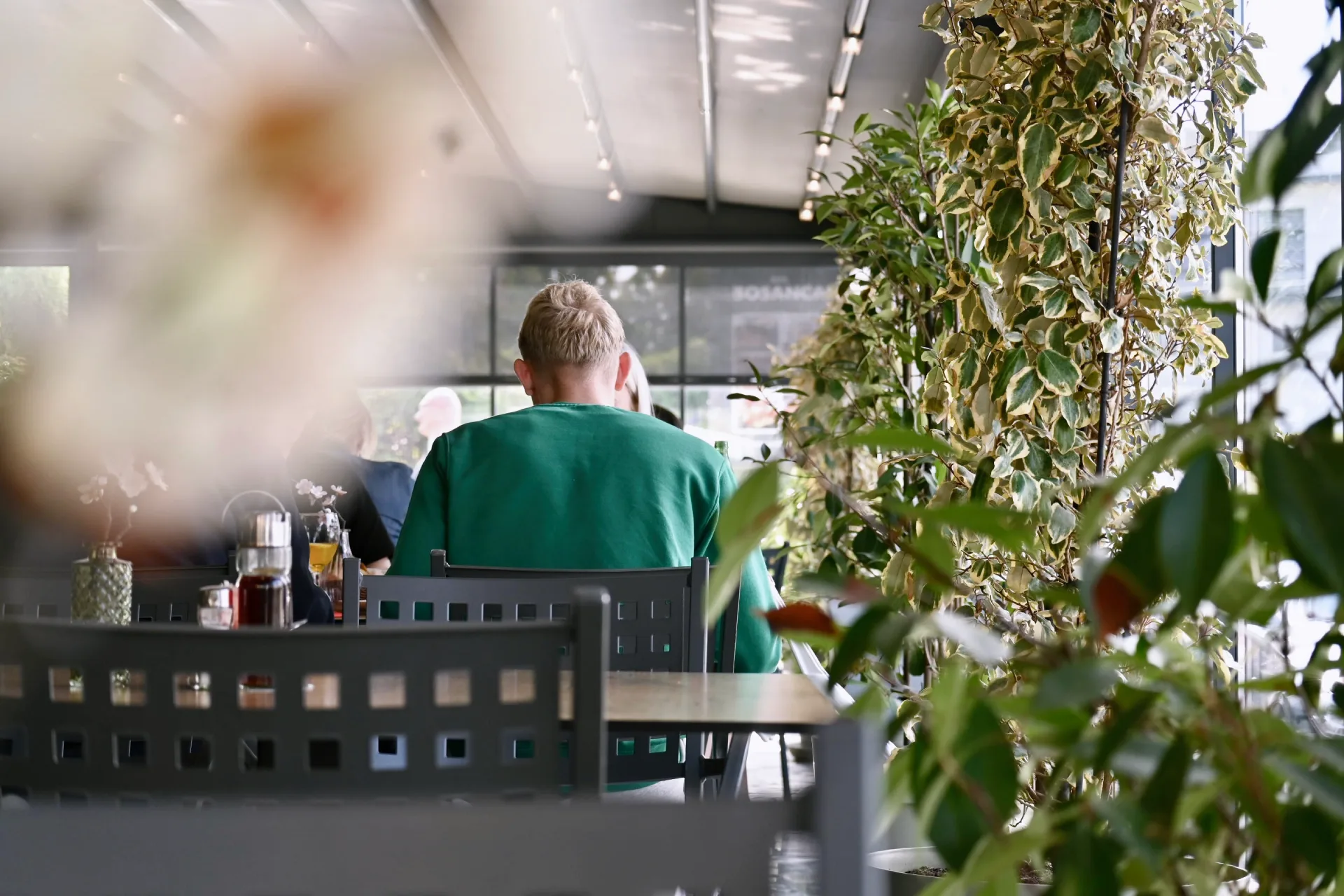 Fotografija crnih stolaca i svijetlosmeđih stolova na terasi dok za jednim stolom sjedi mlađi plavokosi muškarac okrenut leđima