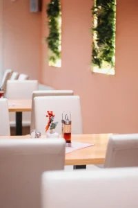 Na slici je prikazana žuto-oker ofarbana prostorija sa svijetim sivkastim stolicama i natur-drvo stolovima a po zidu su zelene florealne strukture